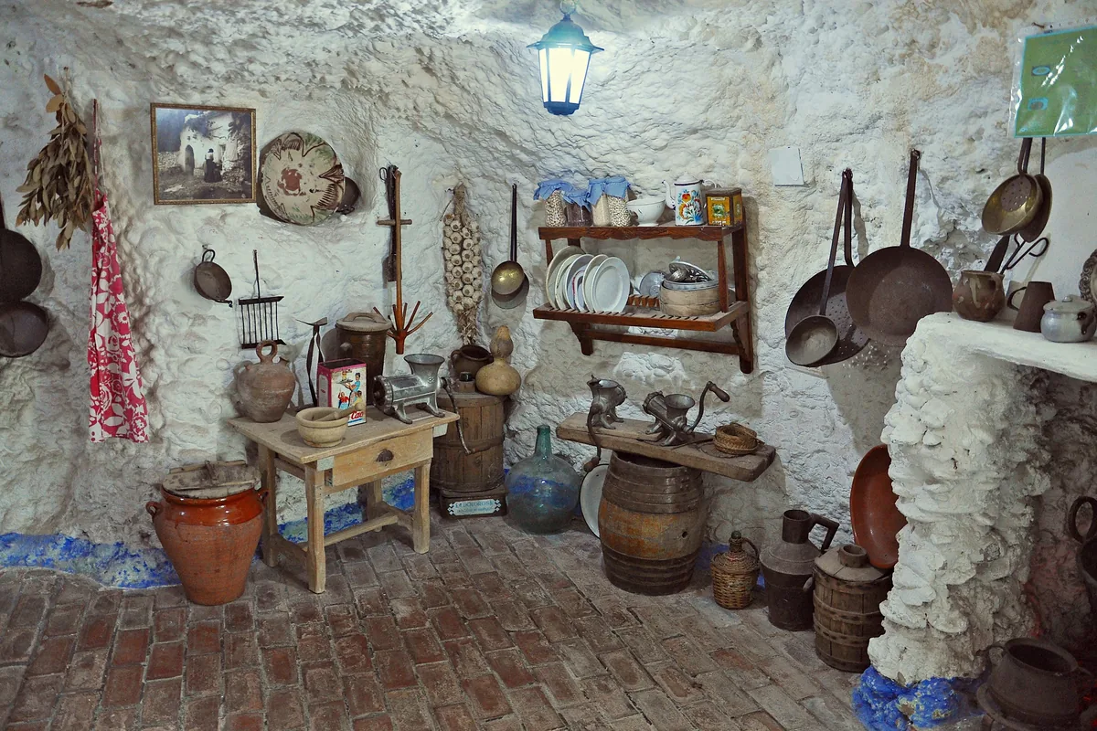 A cave kitchen with rough whitewashed walls, hanging pans, a shelf of plates, a wooden table and a clay jar
