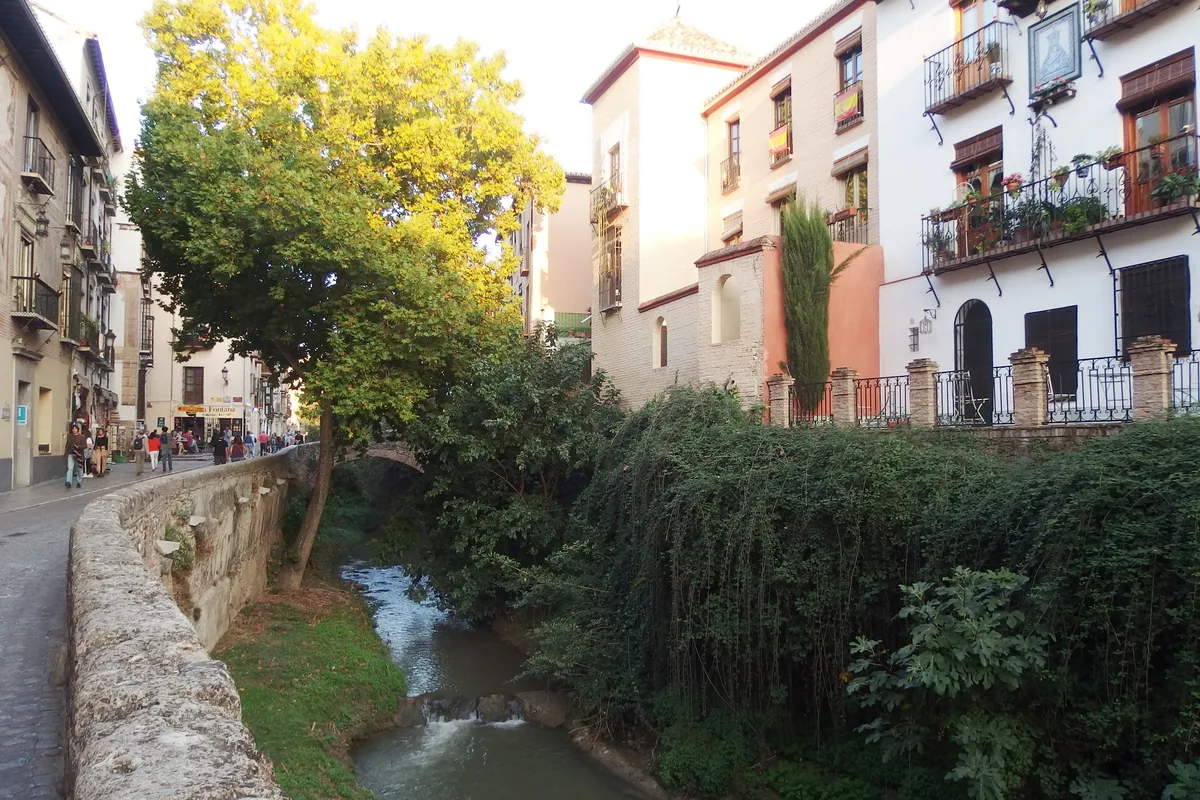 The Darro river running between a stone wall and tall greenery below the white houses of the Albayzín