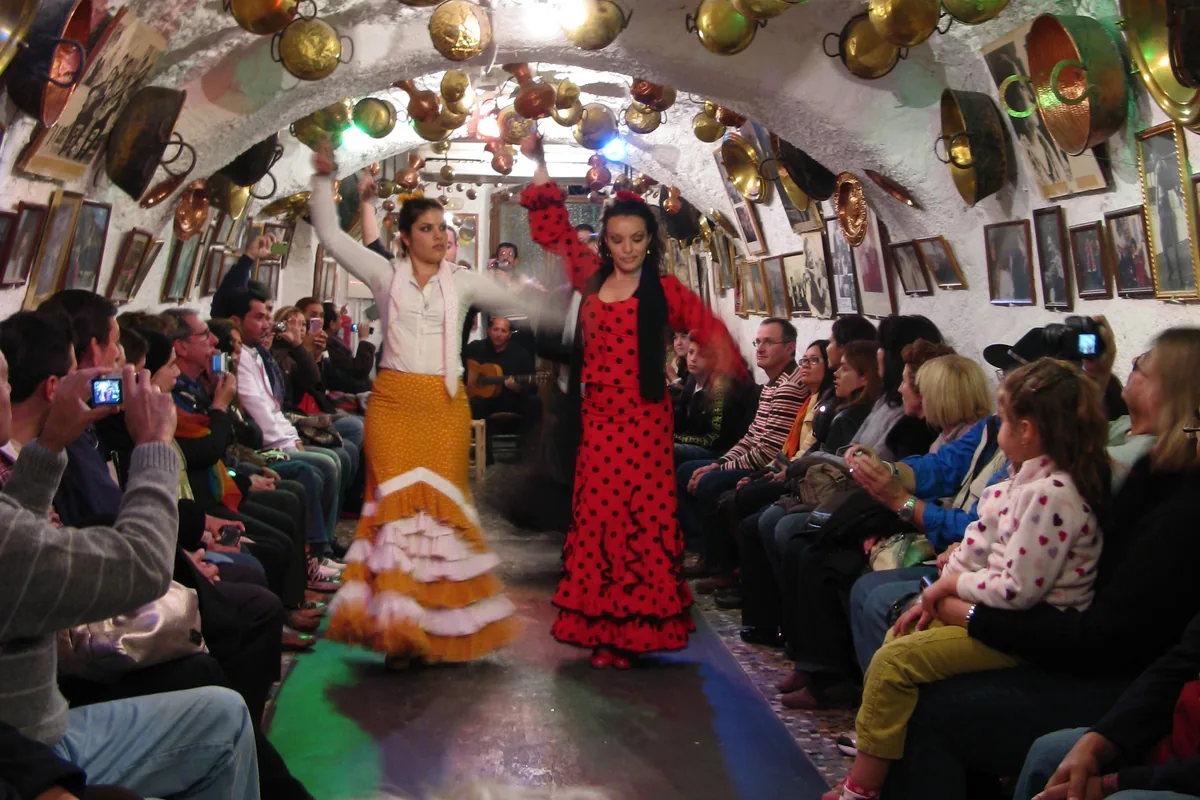 Two flamenco dancers in an orange skirt and a red polka-dot dress under a whitewashed cave arch hung with copper pots, with spectators seated along both walls